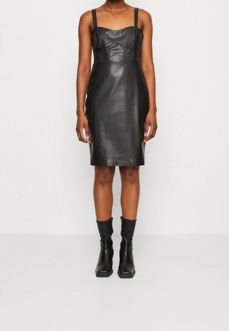 Robe sans manches en similicuir noir avec taille cintrée, ourlet à hauteur des genoux et bottines noires à bout carré avec fermetures éclair latérales.