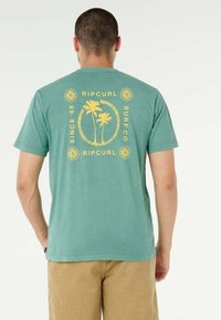 Uomo che indossa una T-shirt verde Ripcurl con grafica di palma gialla e testo "Since '69 Surf Co Ripcurl" sul retro, abbinata a pantaloni beige.