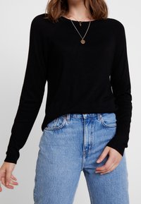Haut noir à manches longues avec manches raglan, associé à un jean bleu clair taille haute. Design simple, texture douce et accents minimalistes.