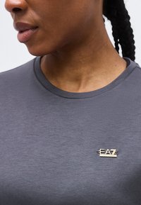 Κοντινό πλάνο ατόμου που φοράει γκρι πουκάμισο με μικρό μεταλλικό λογότυπο "EA7" στο στήθος, πλεξούδες μαλλιών ορατές στο πίσω μέρος.