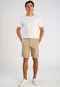 T-shirt blanc à col rond associé à un short beige avec des ourlets retroussés. Porté avec des baskets blanches, se tenant les mains dans les poches.