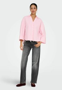 Femme portant une chemise ample rose à boutons, un jean gris droit, une ceinture noire, des ballerines bordeaux et des boucles d'oreilles créoles sur un fond uni.