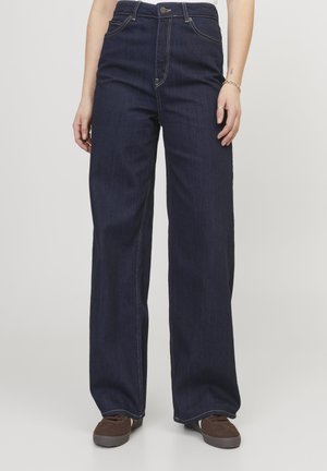 Person wearing high-waisted, dark blue wide-leg jeans paired with brown casual shoes, standing against a plain light background.