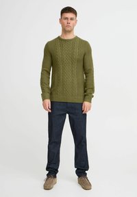 Pulover tricotat în verde măsliniu cu modele texturate de sârmă; guler rotund, manșete și tiv cu nervuri. Asortat cu pantaloni din denim albastru închis și pantofi maro.