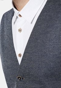 Marinblå texturerad väst med v-ringning, prydd med bruna knappar och en vit skjorta med prickar under. Närbild som visar tyg- och designdetaljer.