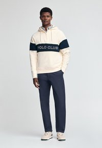 Crème kleurige hoodie met marineblauwe horizontale strepen en het "POLO CLUB" logo. Gepaard met donkerblauwe broek en lichtbeige sneakers met groene accenten.
