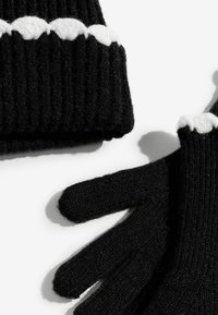 Melnā adīta cepure un piecu pirkstu cimdi ir ar ribotu tekstūru un tiem ir kontrastējoša balta viļņota raksta mala.