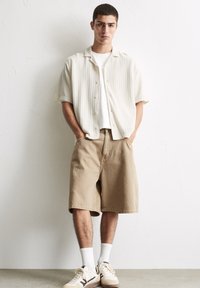 Lysbeige kortærmet skjorte med et struktureret mønster, parret med tan knælange shorts, hvide sokker og cremefarvede sneakers.