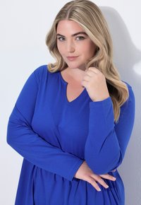 Dlouhé modré šaty s výstřihem do V a nařaseným pasem. Hladká textura, měkká tkanina a uvolněná silueta.