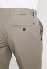 Nahaufnahme der hinteren Tasche einer beigefarbenen Hose mit Knopf und kleinem rot-weiß-blauem Flaggenlogo auf der rechten Seite.