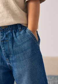 Blugi din denim cu croială lejeră, culoare albastru deschis, textură subtilă și cusături portocalii, prevăzuți cu talie elastică și buzunare laterale.