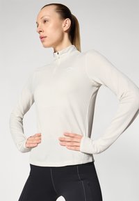 Dugi rukav, kremasti sportski top s polu-zip ovratnikom. Glatka tekstura, uski dizajn, s malim logotipom na prsima i rupama za palčeve u rukavima.