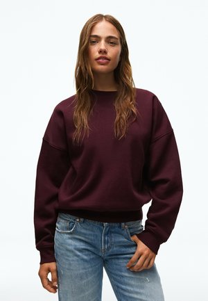 Jeune femme aux longs cheveux ondulés, portant un sweat-shirt bordeaux foncé et un jean bleu, debout devant un fond blanc uni.