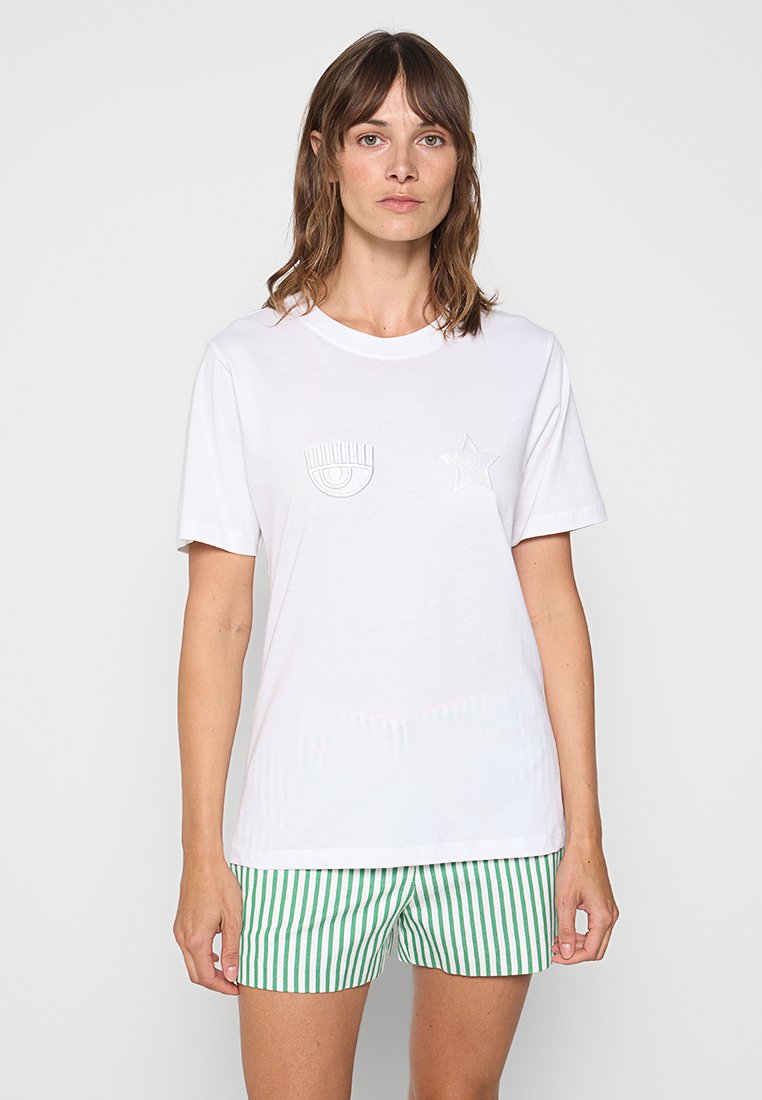 T-shirt en coton blanc à col rond, avec des motifs brodés d'œil et d'étoile. Le modèle porte un short à rayures vertes et blanches.
