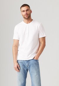 Witte korte mouwen henley shirt met drie knopen, gedragen met lichtblauwe denim jeans. De stof lijkt zacht; het shirt heeft een subtiele logo-detail.