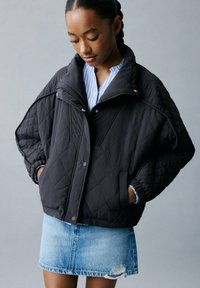 Veste matelassée noire avec un col montant et une fermeture éclair à l'avant. Présente des poches latérales et des poignets élastiqués. Portée par-dessus une chemise à rayures bleues et une jupe en denim.