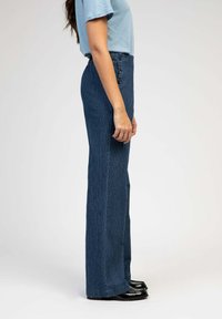 Pantalons taille haute, bleu foncé à rayures, avec des boutons sur les côtés, jambes évasées et une texture lisse, associés à des chaussures noires.