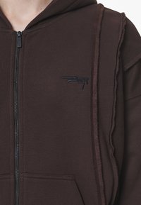 Sudadera con cremallera marrón hecha de un tejido suave con un diseño en capas. Cuenta con capucha, bolsillos laterales y un logo bordado en el pecho.