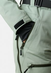 Zielona kurtka typu soft shell z czarną wewnętrzną warstwą, kieszeniami na zamek i zapięciem na rzep; charakteryzuje się smukłym kształtem i teksturowanym materiałem.