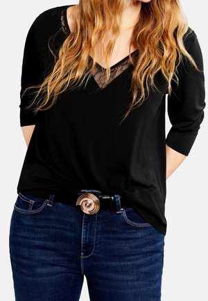 Femme aux longs cheveux ondulés portant un top noir avec un détail en dentelle et un jean bleu, accessoirisée d'une ceinture avec une grande boucle en spirale.
