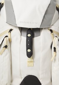 Reppu, jossa on harmaa verkko päällinen, beige kangas, useita vetoketjullisia taskuja, mustia yksityiskohtia ja näkyvä Oakley-logo etupuolella.
