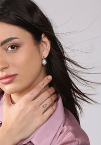 Silberne Creolen mit einem Perlenanhänger, strukturierten Akzenten und einem Modell, das eine pinke Satinbluse trägt, während sie Tätowierungen an ihrer Hand zeigt.