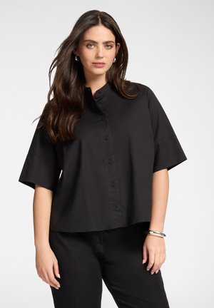 Kobieta z długimi ciemnymi włosami, ubrana w czarną koszulę z krótkim rękawem zapinaną na guziki oraz czarne spodnie, stojąca na jednolitym jasnym tle.