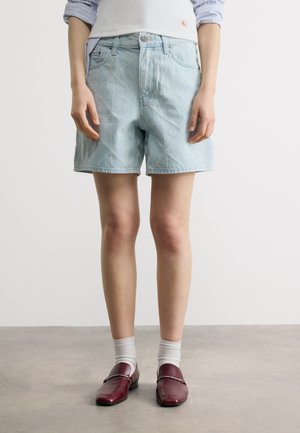 Személy világoskék farmer rövidnadrágot visel enyhe mintával, fehér zoknival és bordó bőrből készült, láncdíszes kényelmes cipővel.