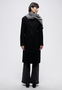 Fekete hosszú kabát testhez simuló szabással, kihajtós gallérral és két elülső gombbal. Szürke sállal és bő szárú fekete nadrággal párosítva.