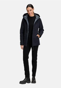 Veste imperméable bleu marine avec doublure grise, capuche ajustable, poches zippées et coupe légèrement cintrée, associée à un pantalon noir et des bottes.