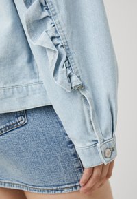 Chaqueta de mezclilla de color azul claro con detalles de volantes en las mangas, con puños abotonados y una textura suave, combinada con una falda de mezclilla.
