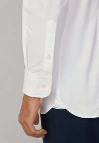 Camicia bianca con colletto a punta, caratterizzata da un singolo bottone e una texture sottile. Tessuto liscio con orlo arrotondato e leggero lucido.