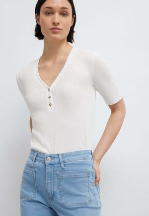 Donna con capelli corti e scuri che indossa una maglietta bianca a maniche corte con scollo a V e bottoni e jeans azzurri a vita alta, in piedi contro uno sfondo semplice.