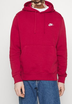 Person trägt einen roten Nike-Hoodie mit Fronttasche und blaue Jeans, steht vor einem einfarbigen, hellen Hintergrund.