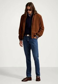 Chaqueta bomber de ante marrón sobre un suéter navy, combinada con jeans de mezclilla azules y mocasines marrones, complementada con gafas de sol oscuras y un reloj.