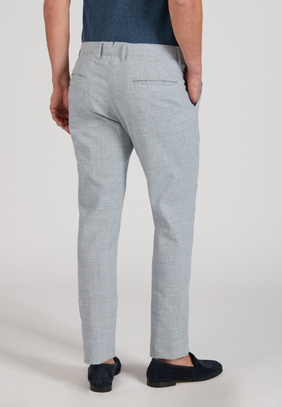 Man wearing light gray checkered trousers and dark blue loafers, standing with one hand in pocket, shown from the back.