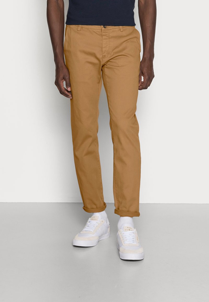 Kaki chinos med rak passform, tillverkade av bomull, med knappstängning och upprullade ärmar, parat med vita sneakers och strumpor.