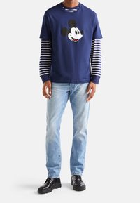 Námořnické tričko s grafikou Mickey Mouse, vrstvené přes šedé a bílé pruhované dlouhé rukávy. Světle modré džíny a černé boty dokončují celkový vzhled.