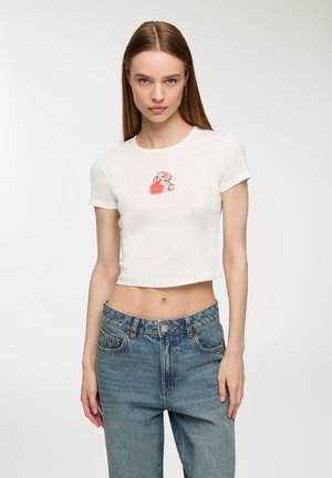 T-shirt bianco corto con maniche corte, caratterizzato da una grafica stampata di un cane su un cuore rosso. Abbinato a jeans denim blu a vita alta.