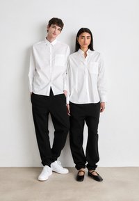 To personer står side om side, iført hvite skjorter med brystlommer og svarte, vide bukser. De har på seg hvite sneakers og svarte leiligheter.