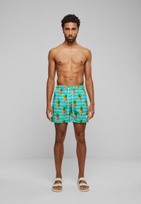 Shorts de bain avec un fond turquoise, présentant un motif d'ananas orange et des accents géométriques. Accompagnés de sandales blanches.