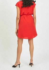 Robe rouge à manches courtes en tissu léger, elle présente une taille ceinturée, une longueur au-dessus du genou et une texture lisse et transparente. Portée avec des escarpins noirs.