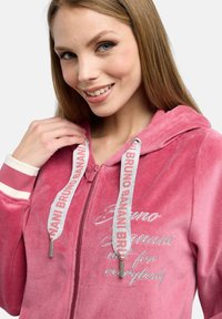 Rosa sammetshoodie med dragkedja framtill, broderad text och snoddar som har en repetitiv logotyp. Mjuk textur och avslappnad design.
