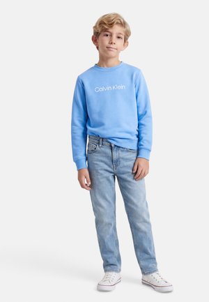 Băiat stând cu mâna relaxată pe lângă corp, purtând un hanorac Calvin Klein albastru deschis, blugi albastru deschis și adidași albi, pe fundal alb.