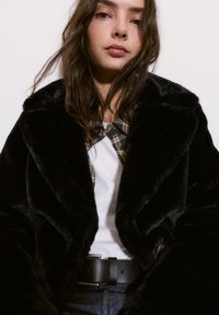 Czarny płaszcz z futra alternatywnego z szerokimi klapami, noszony na białej koszuli i w kratkę. posiada czarny pasek i fakturowany materiał.