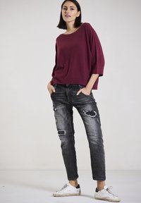 Burgunderfarbener locker gestrickter Pullover kombiniert mit zerrissenen schwarzen Jeans. Das Model trägt weiße Sneakers mit schwarzen Akzenten. Glatte Textur und lässiges Design.