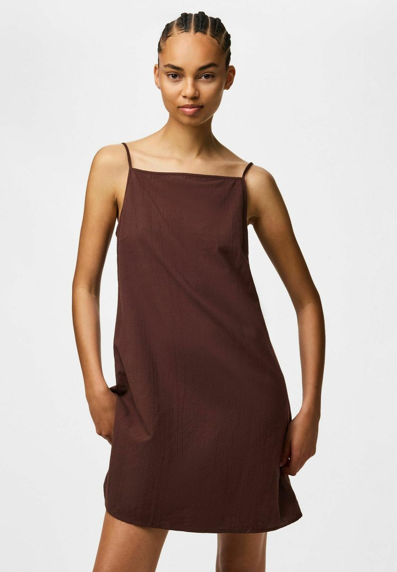 Jeune femme aux cheveux tressés portant une robe marron sans manches à fines bretelles, posant les mains dans les poches devant un fond uni.