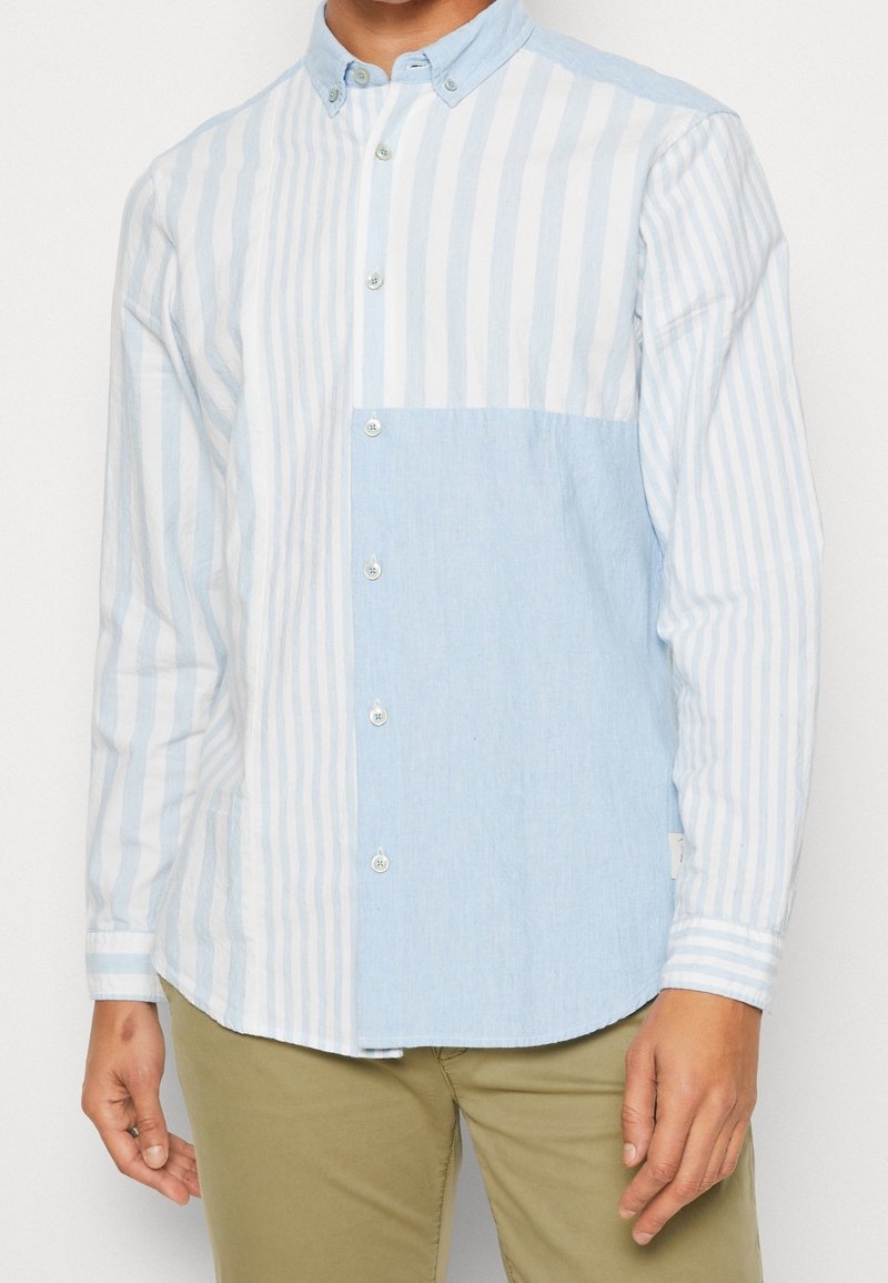 Camisa de botones de rayas azul claro y blanca con un panel sólido azul claro en contraste, hecha de una tela texturizada, que presenta un cuello abotonado.