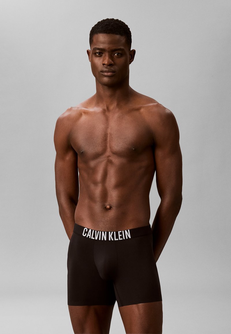 Homme musclé de face portant un boxer noir Calvin Klein sur un fond gris uni.