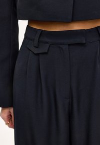 Pantalon sur mesure bleu marine avec une ceinture plate, des plis à l'avant, et un accent rectangulaire sur la boucle de ceinture. Texture tissée lisse.